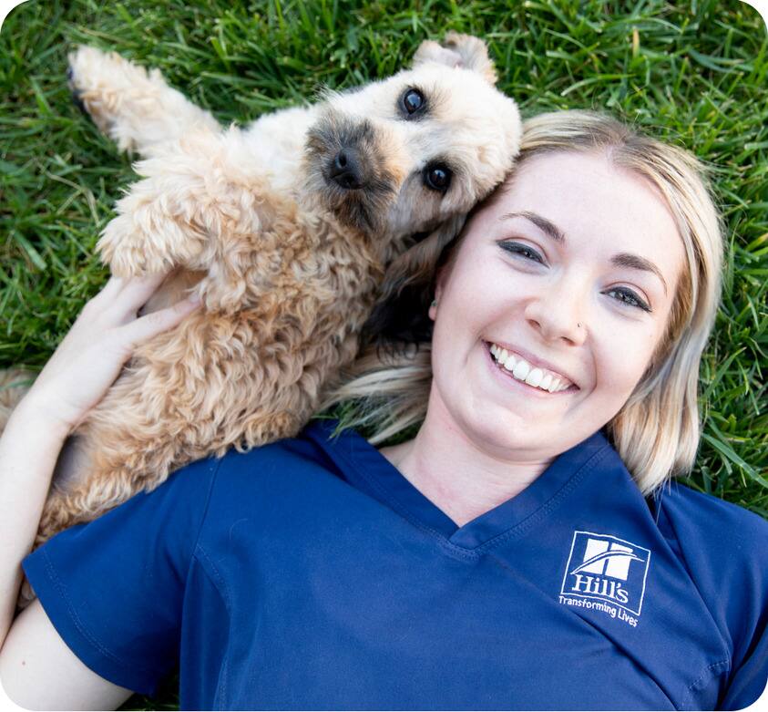Un perro pequeño acostado en el césped junto a una investigadora de Hill's