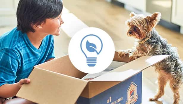  Un niño pequeño y un perro mirando una caja reciclable de Hill’s con un ícono de reducción de desperdicios